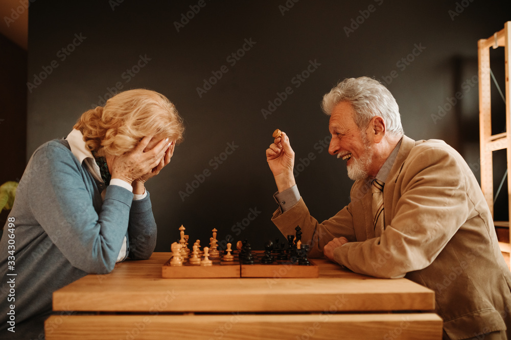 Close photo of elderly couple playing chess at home. Woman is not happy because man is winning.