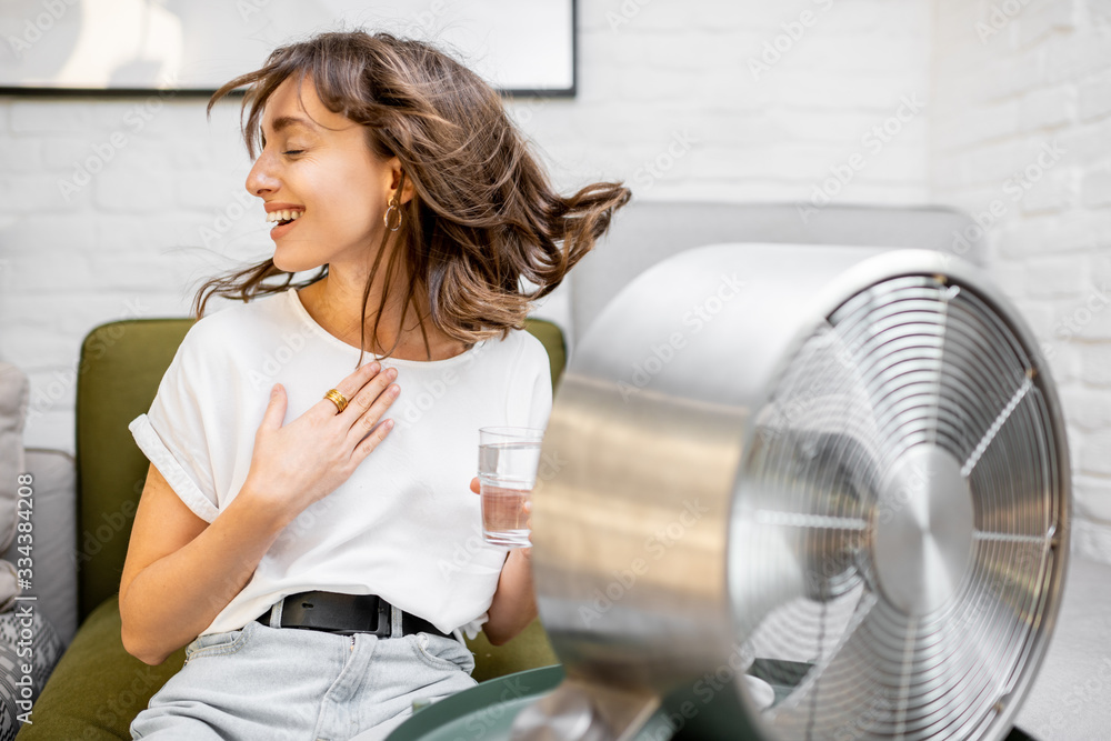 Young woman feeling hot, enjoying the flow of air from the fan, sitting on the couch with a glass of