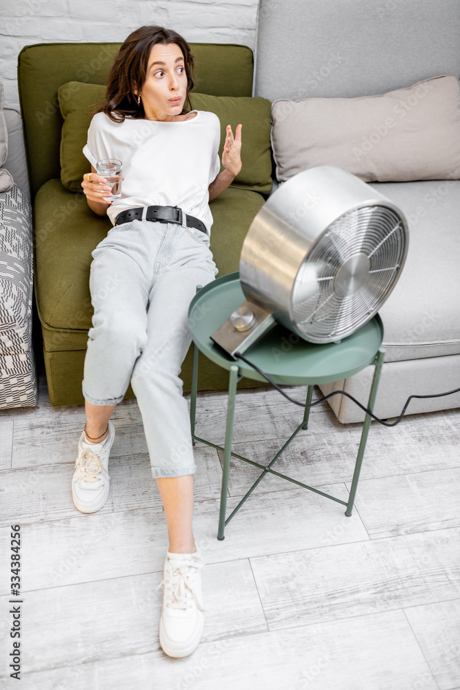 Young woman feeling hot, enjoying the flow of air from the fan, sitting on the couch with a glass of