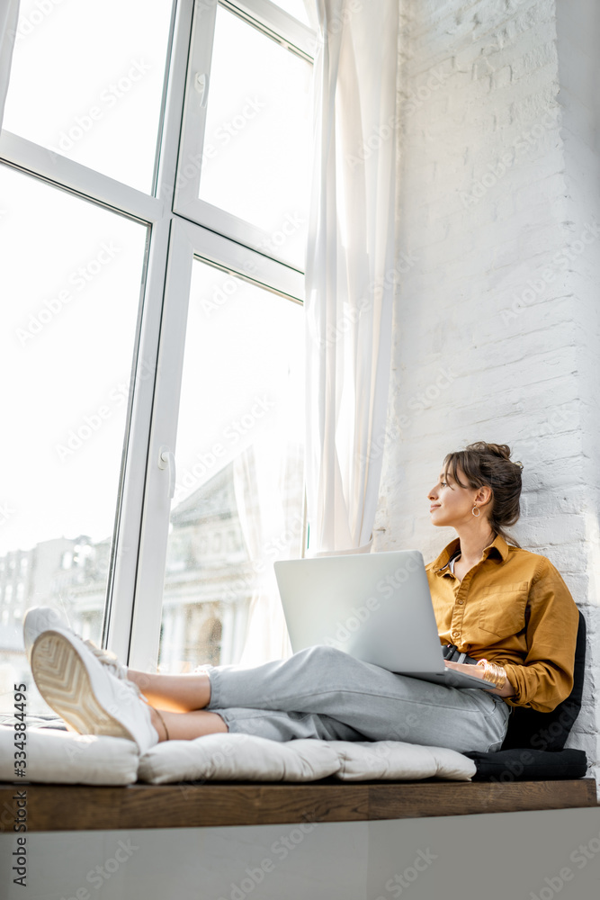 Young woman dressed casually working on laptop while sitting on the window sill at home. Work from h