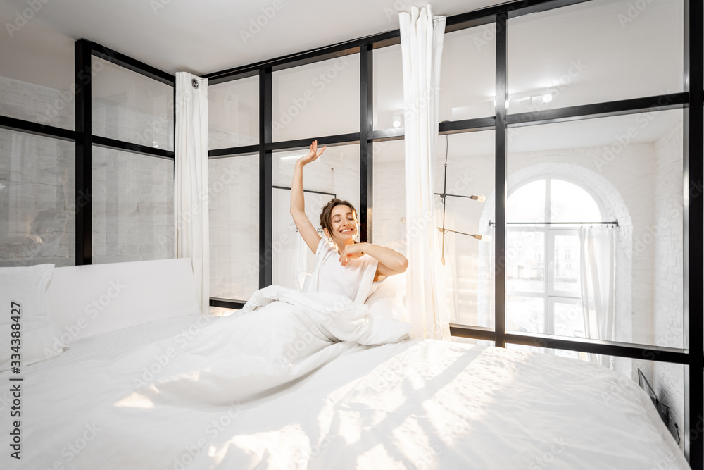 Young woman waking up in the beautiful white and sunny bedroom, wide interior view. Good morning con