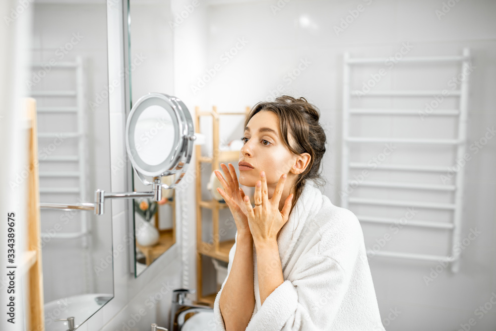 Young woman in bathrobe taking care of her skin, looking at the mirror in the bathroom. Facial skin 