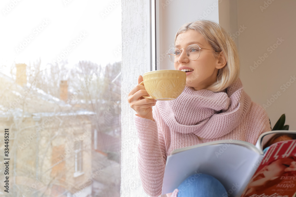 Beautiful young woman drinking tea and reading magazine near window at home