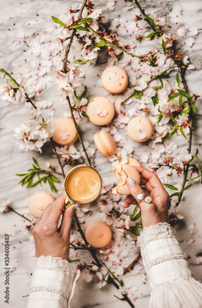 Flat-lay of womans hands holding sweet macaron cookies and fresh coffee in cup over white spring blo