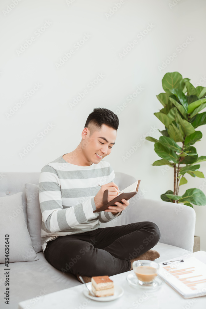 Asian man writing on notebook when sitting on sofa at home.