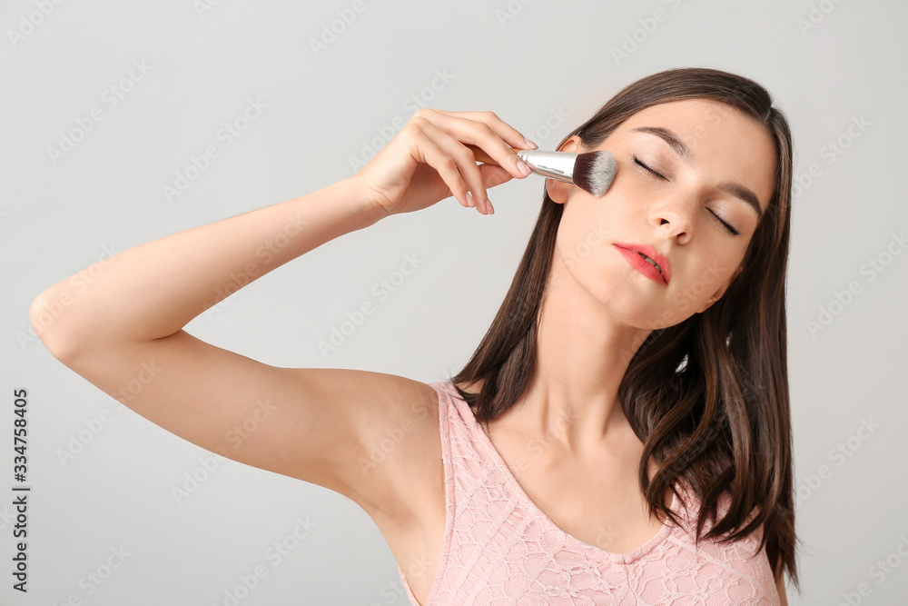 Beautiful young woman applying makeup on light background