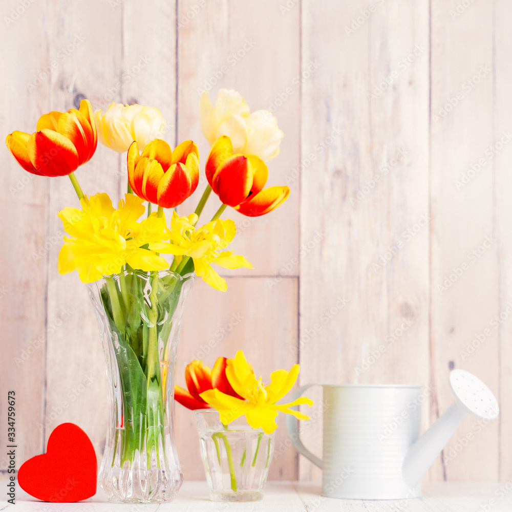 Tulip flower arrangement in glass vase with heart greeting, watering can decor on wooden table backg