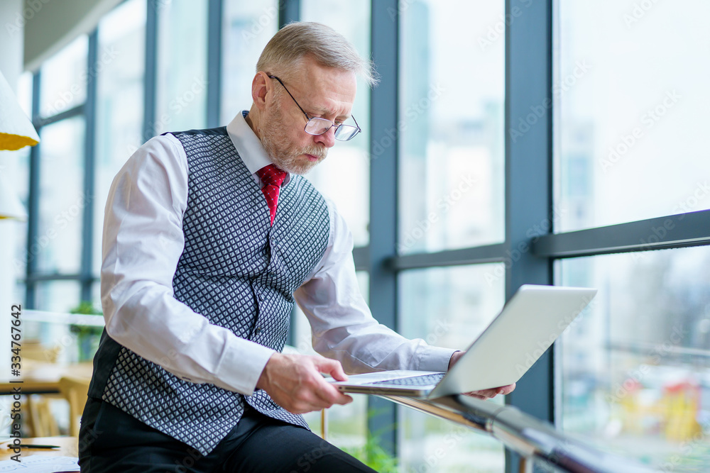 Mature businessman looking at laptop reading printed documents. Sitting near the window in office. P