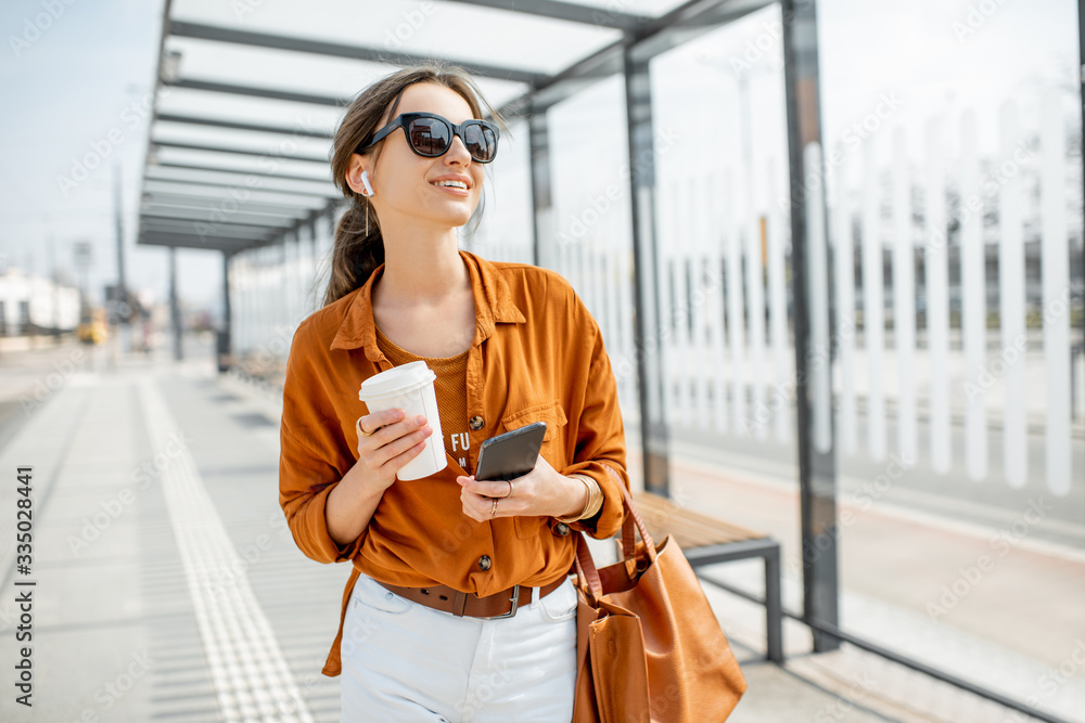 Lifestyle portrait of a young and cheerful woman standing with phone and coffee cup on the public tr