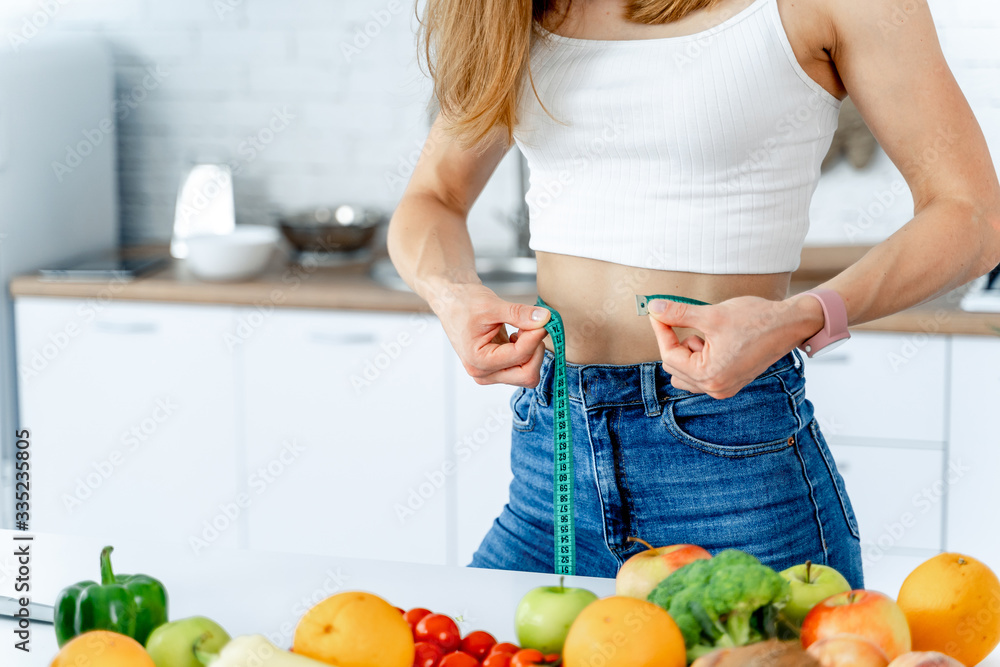 饮食理念。厨房里有一位漂亮的年轻女士，手里拿着健康的食物。腰部有卷尺。Fr