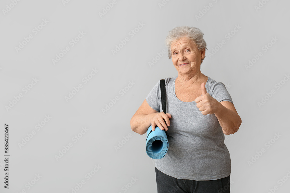 Elderly woman with yoga mat showing thumb-up gesture on light background