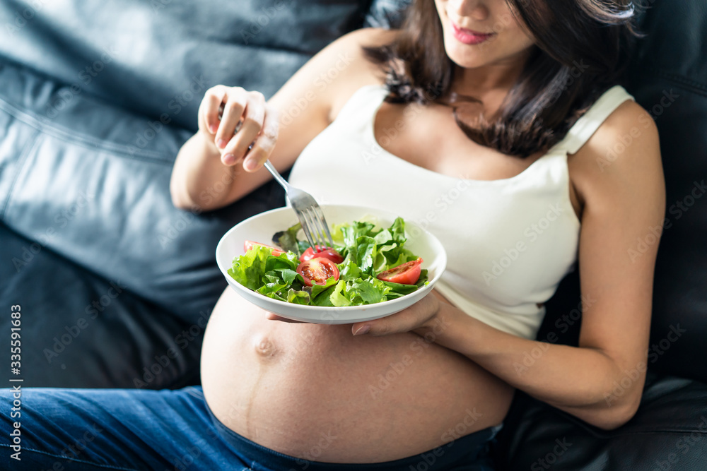 Pregnant women sitting on sofa is holding salad bowl in her left hand. She is holding fork with red 