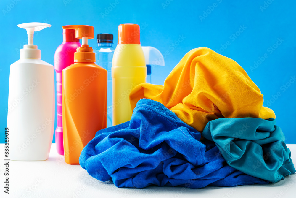 Plastic bottles of cleaning products set with pile clothes on white table blue background.