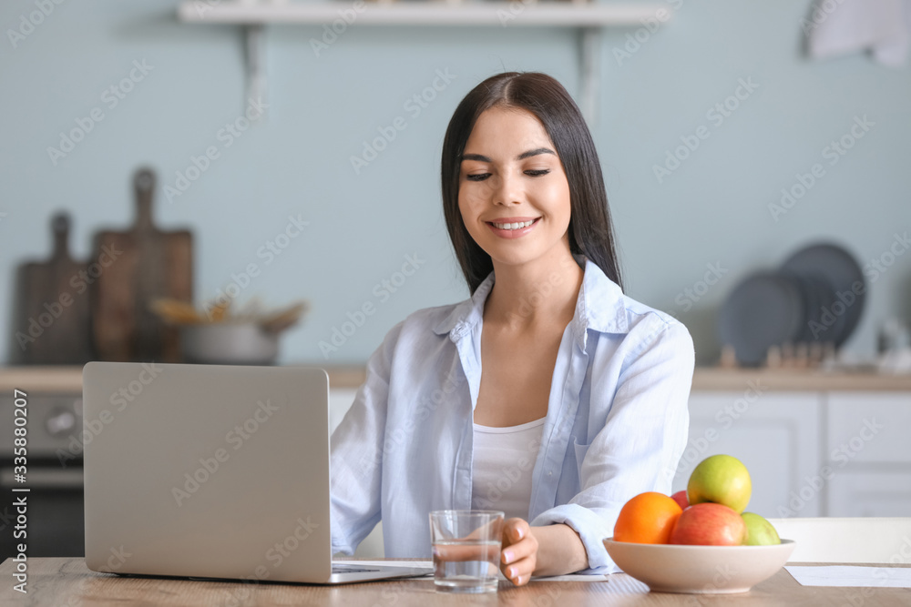 Beautiful young woman with laptop drinking water in kitchen