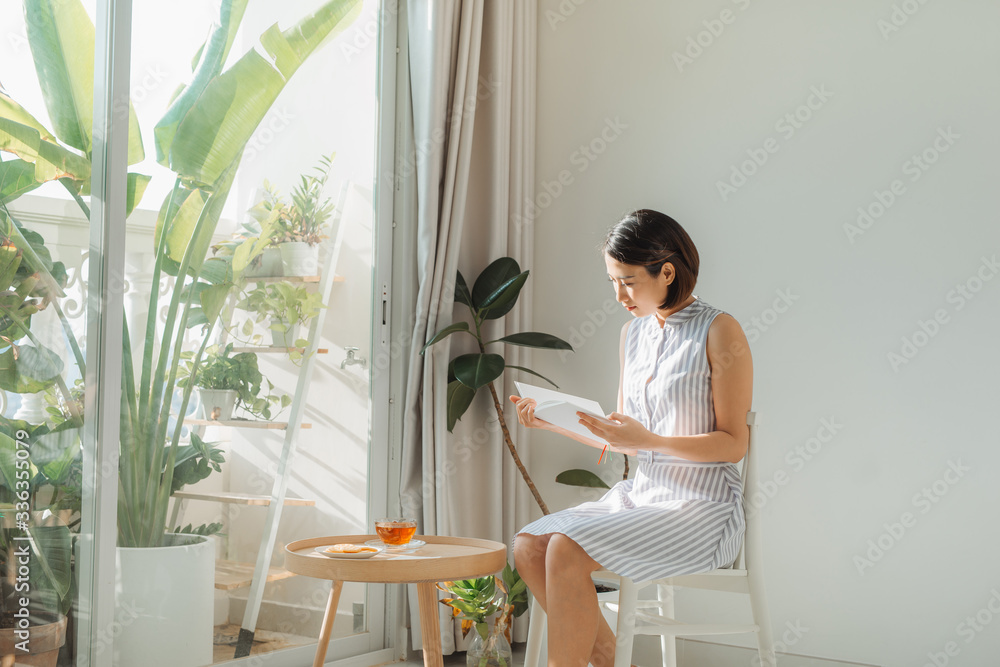 年轻迷人的女人坐在椅子上，在家里的窗户旁边看书。