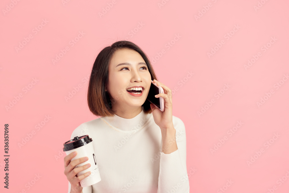 Portrait of cheerful young beautiful Asian woman listening smartphone while holding coffee cup over 