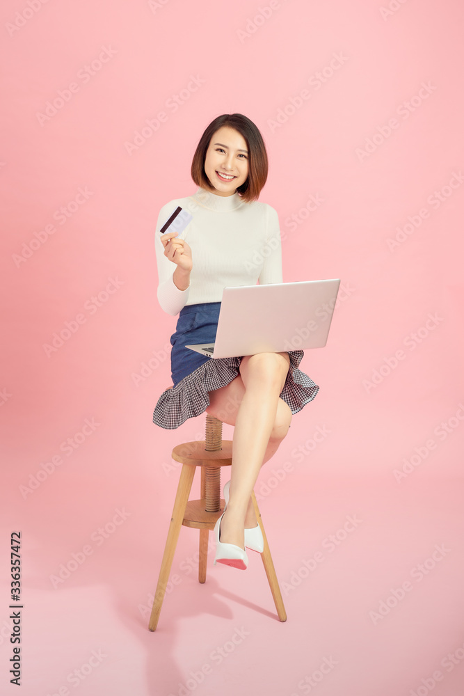 Young Asian woman using credit card to payment online with her laptop. Isolated on pink background.