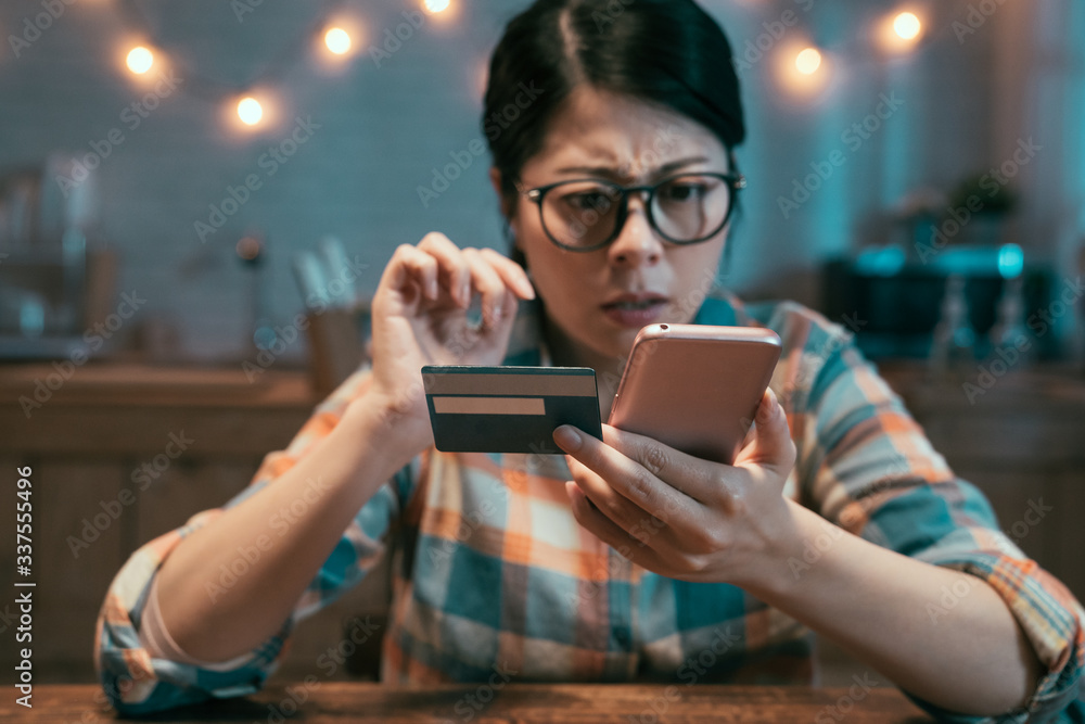 Confused asian chinese woman paying online with credit card sitting in dark home kitchen at night. s