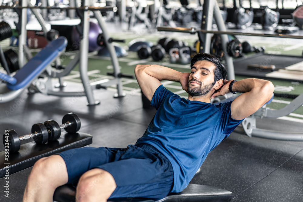 Young fit handsome smiling Caucasian man exercising by doing a sit-up on bench in gym or fitness clu