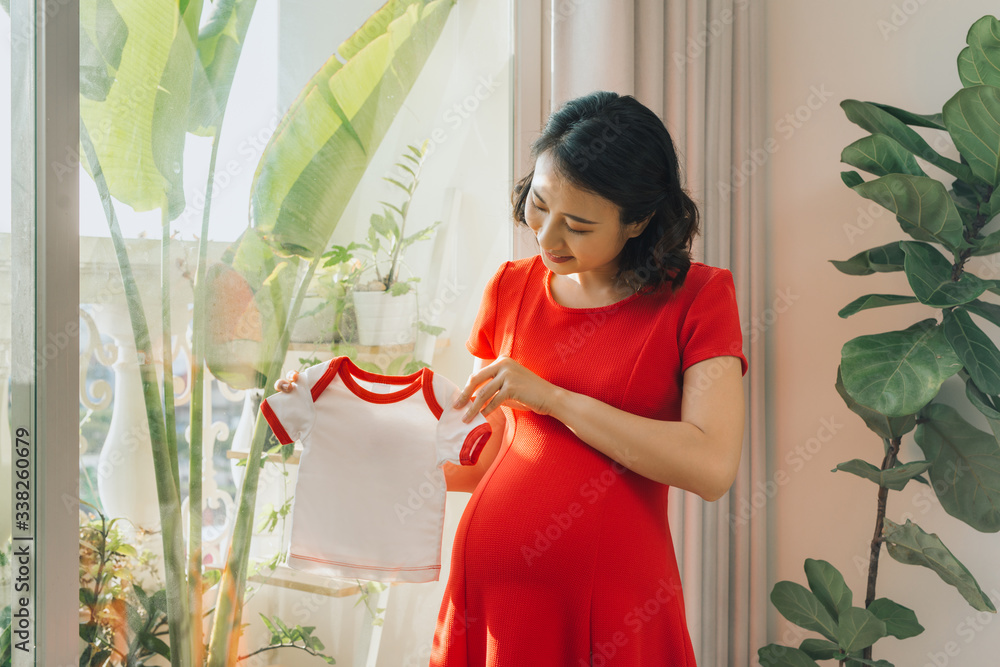 年轻的孕妇穿着她未来婴儿的衣服站在大窗户附近