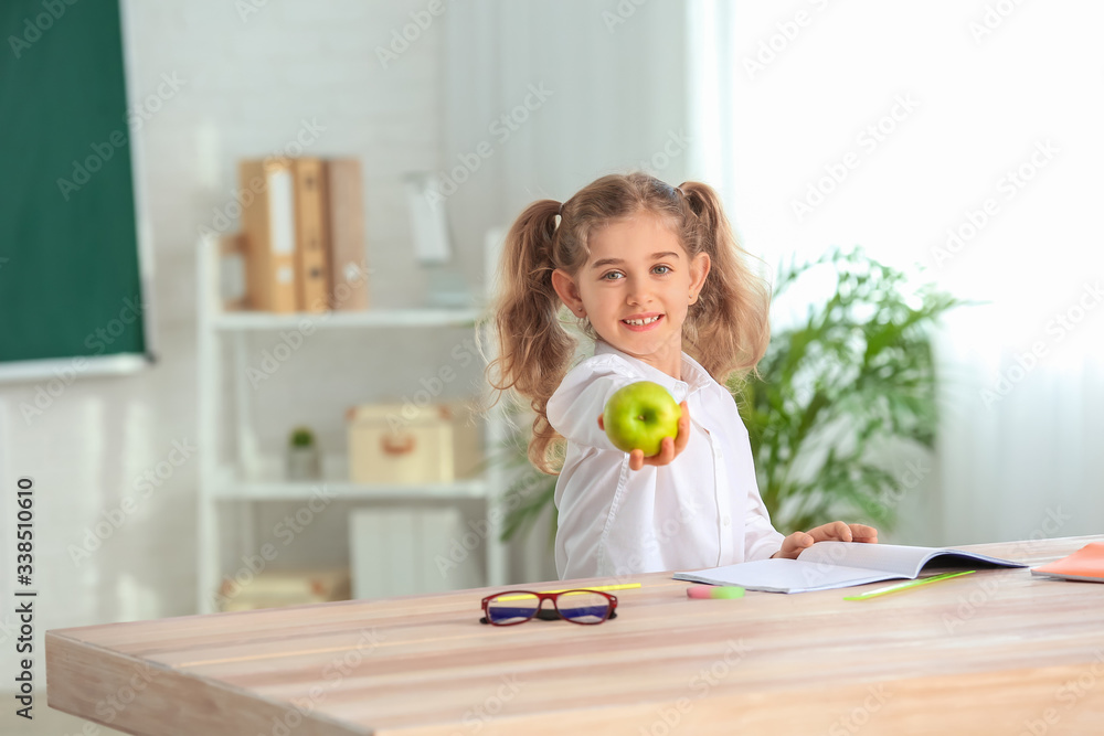 Cute little schoolgirl with apple at desk in classroom