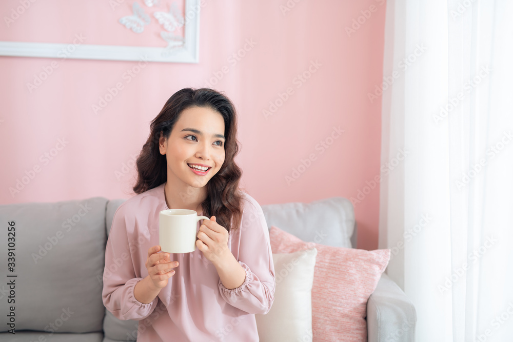 Cheerful young Asian woman holding a mug when sitting on sofa at home.