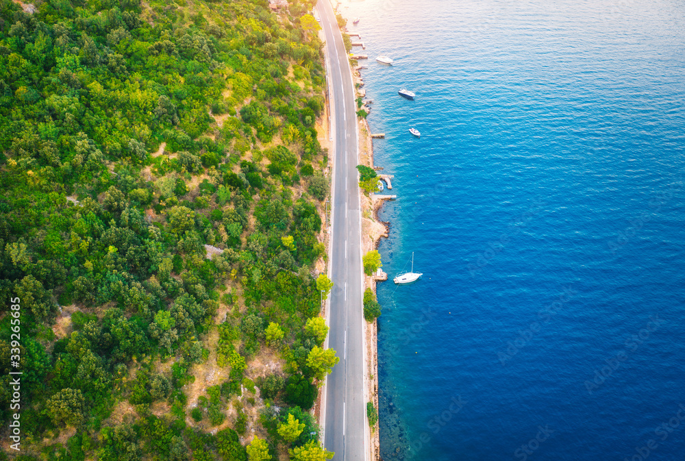 夏日日落时，美丽的绿色森林中的道路和大海中的船只的鸟瞰图。五颜六色的蓝