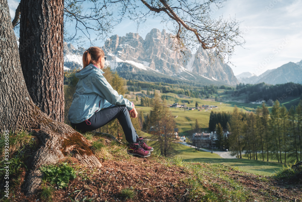 Beautiful young woman in sunglasses and blue jacket is sitting on the hill under the tree at sunset.