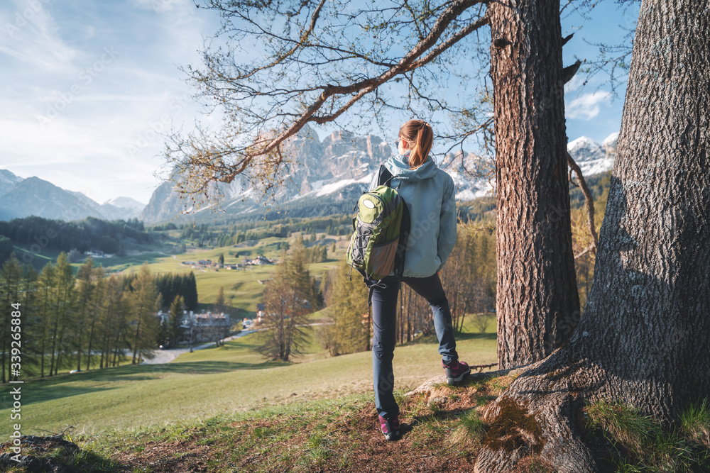 Beautiful young woman in sunglasses and blue jacket is standing on the hill under the tree at sunset