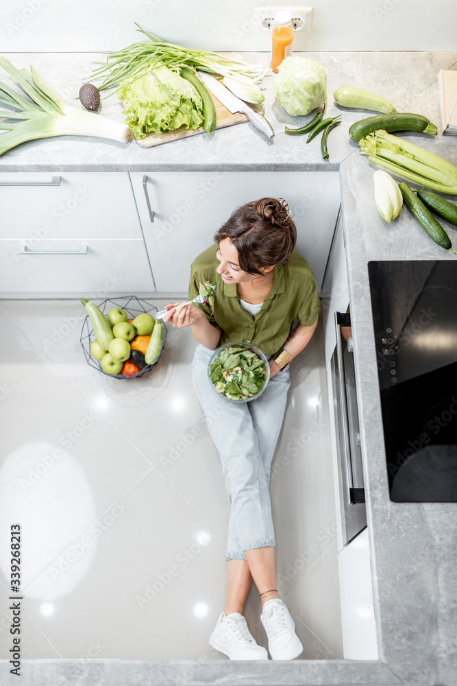 一位女士坐在厨房地板上，靠近装满新鲜食材的桌子，一边吃着健康的沙拉