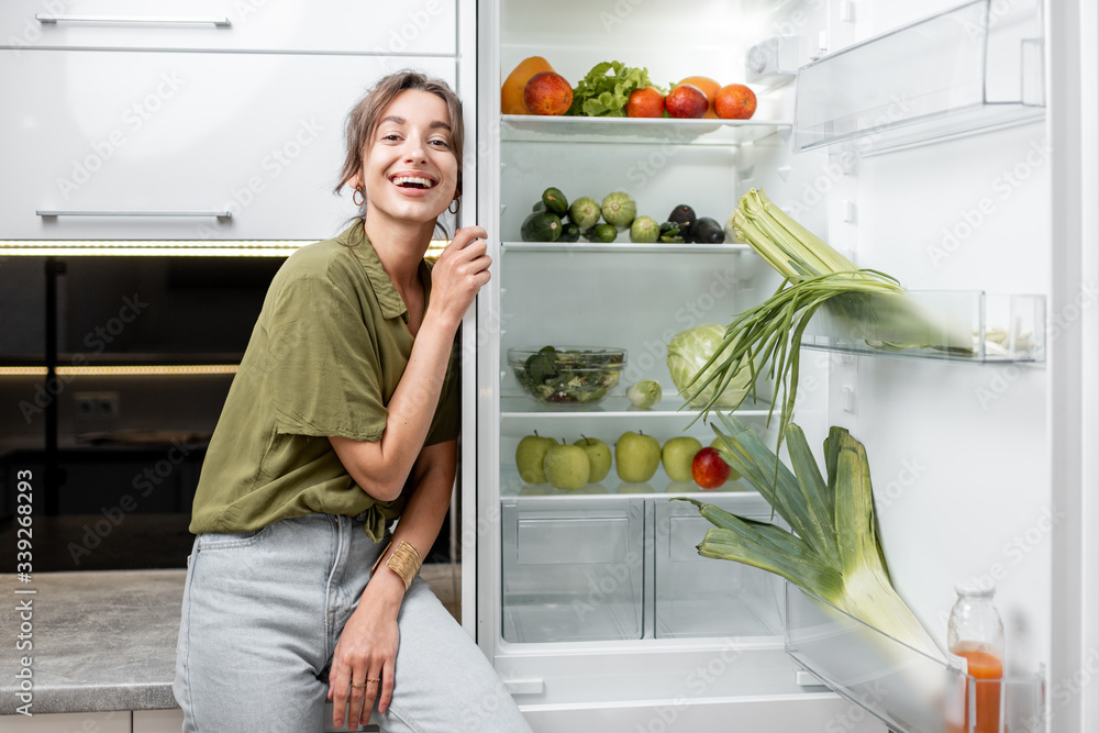 Portrait of a young woman sitting on the kitchen table near the fridge full of fresh and healthy foo