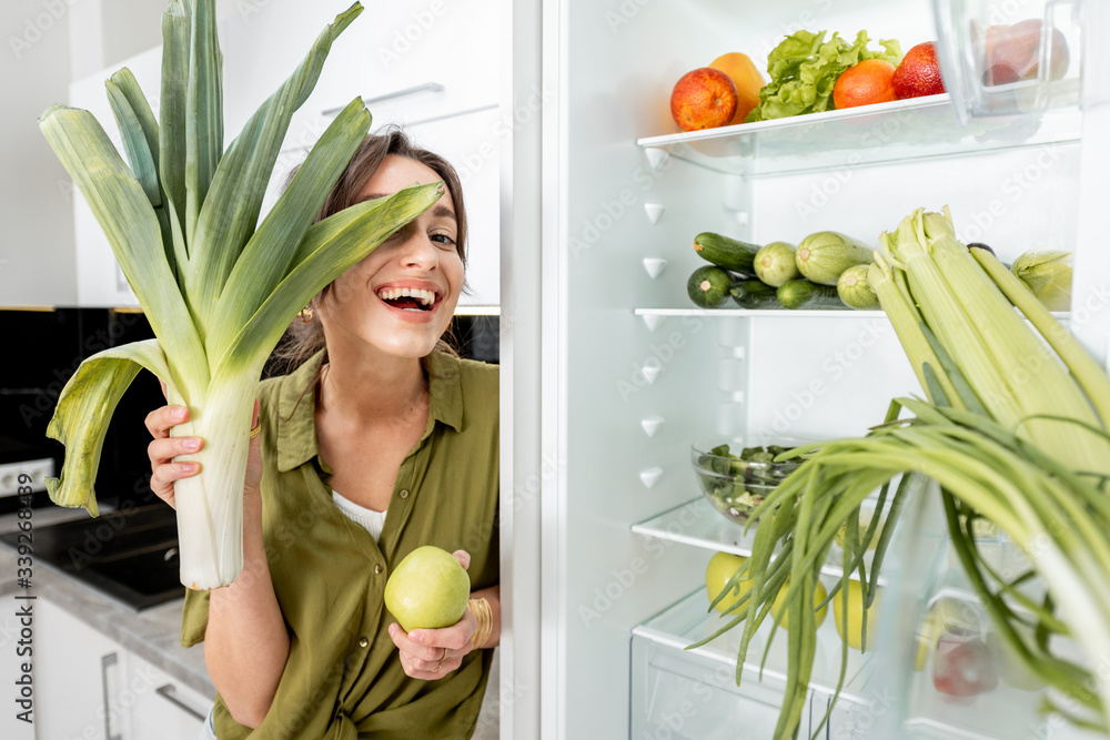 Portrait of a young and joyful woman with fresh greens near the fridge on the kitchen at home. Healt