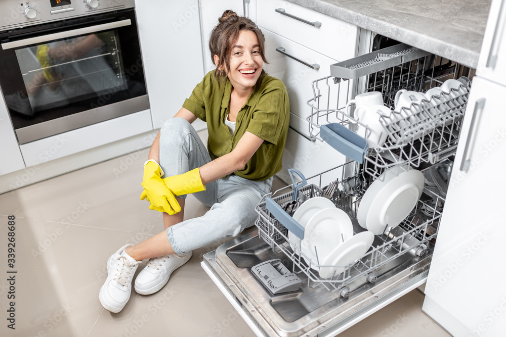 Portrait of a happy housewife sitting near the dishwasher with clean dishes on the kitchen at home. 