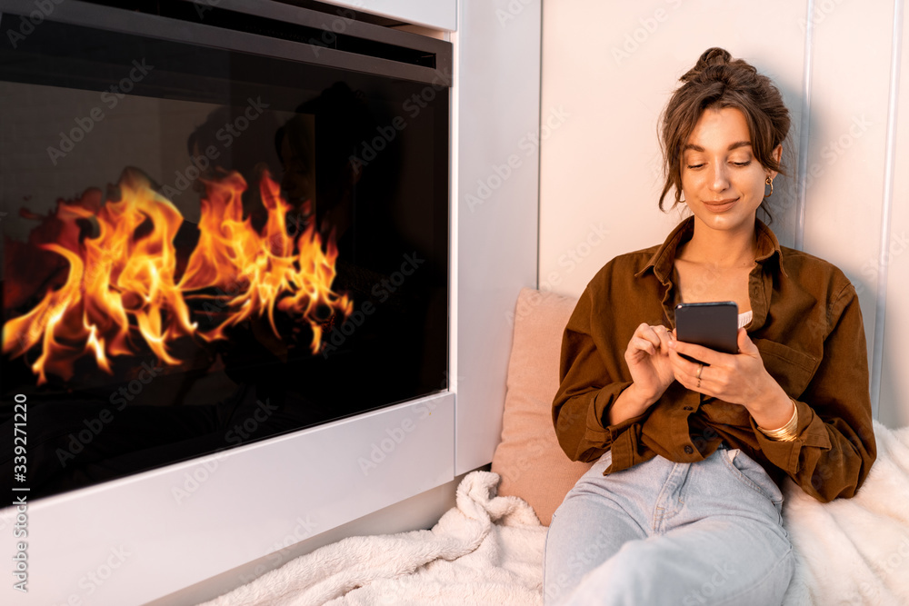 Young woman relaxing with a smart phone, sitting near the fireplace at the modern living room at hom