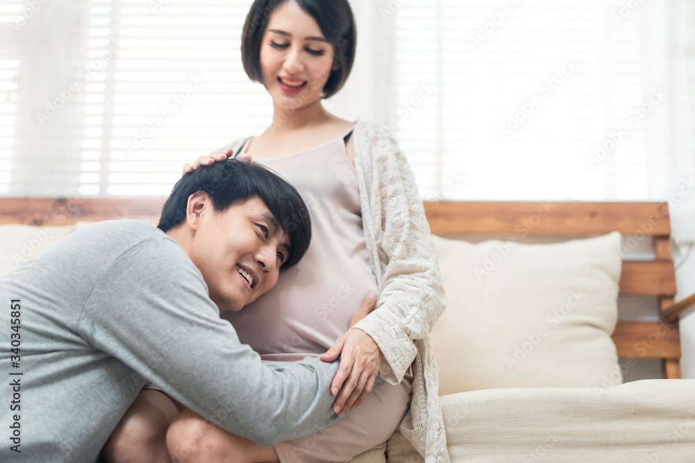 Asian man and pregnancy woman sit on sofa at home. Father holding mother tummy listen and hear infan