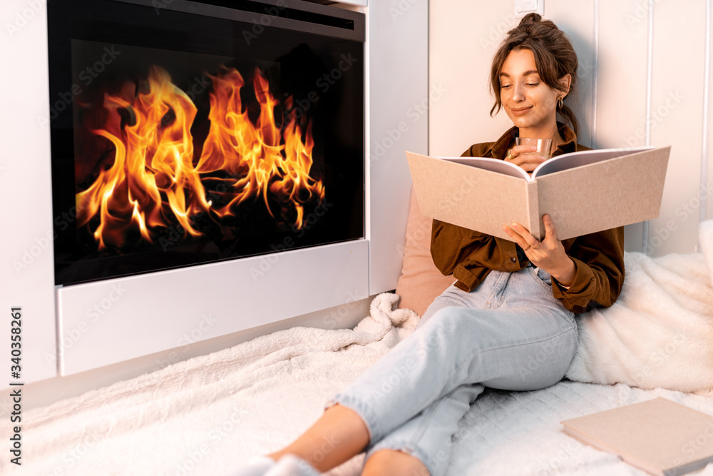 Young woman reading book, relaxing near the fireplace at the modern living room at home