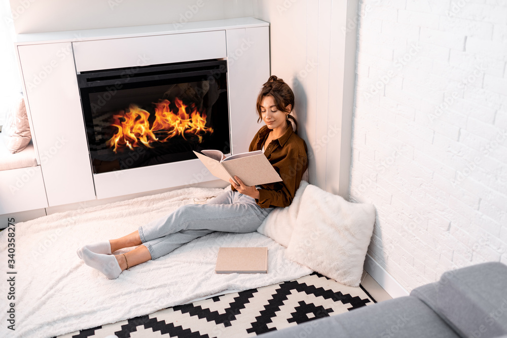 Young woman reading book, relaxing near the fireplace at the modern living room at home