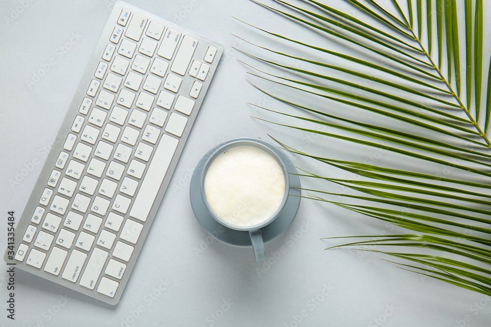 Cup of coffee, PC keyboard and tropical leaf on light background