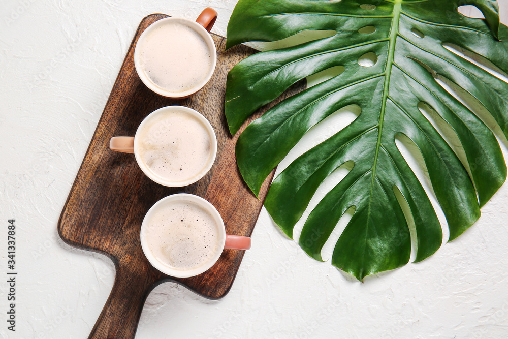 Board with cups of coffee and tropical leaf on light background