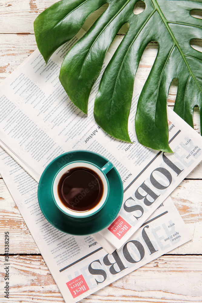 Cup of coffee, newspaper and tropical leaf on light wooden background