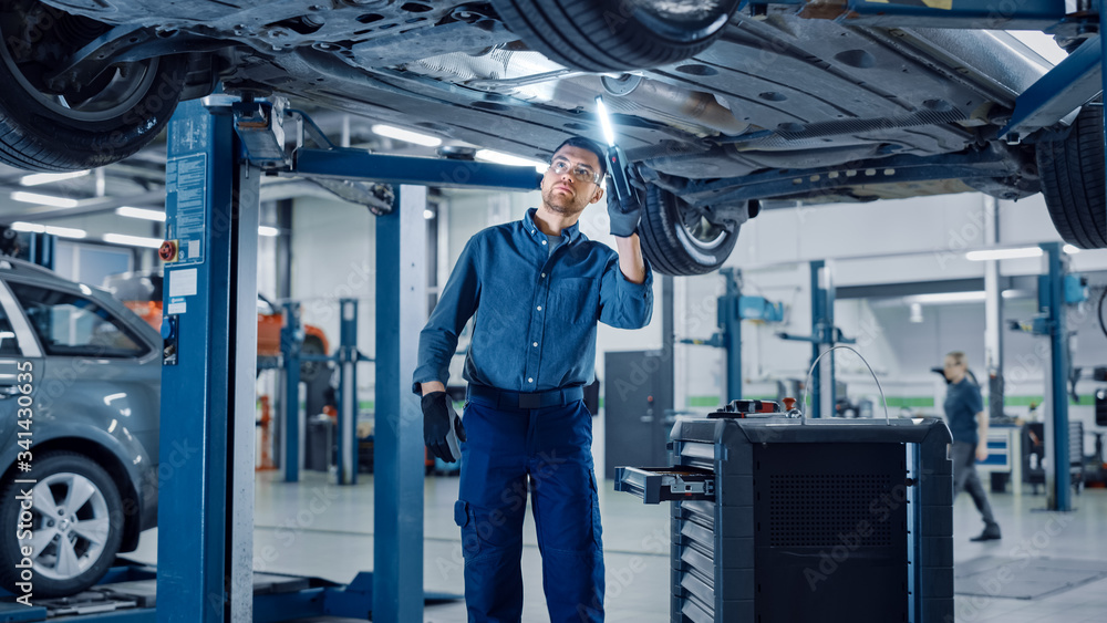 Handsome Professional Car Mechanic is Working Under a Vehicle on a Lift in Service. Repairman is Usi