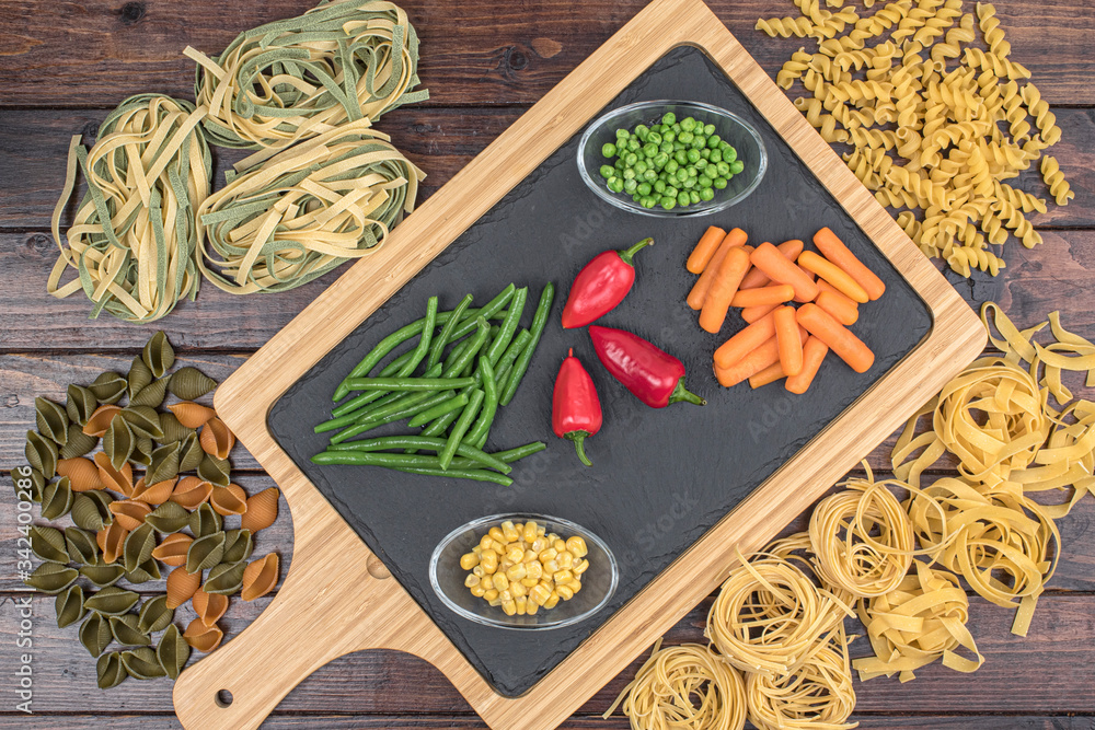 Dried mixed pasta and vegetables on cutting board on the dark wooden table.Top view.