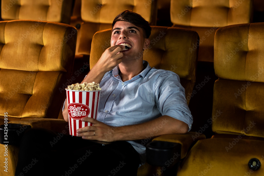 A young man smiling enjoying with his popcorn while watching a movie in theater cinema