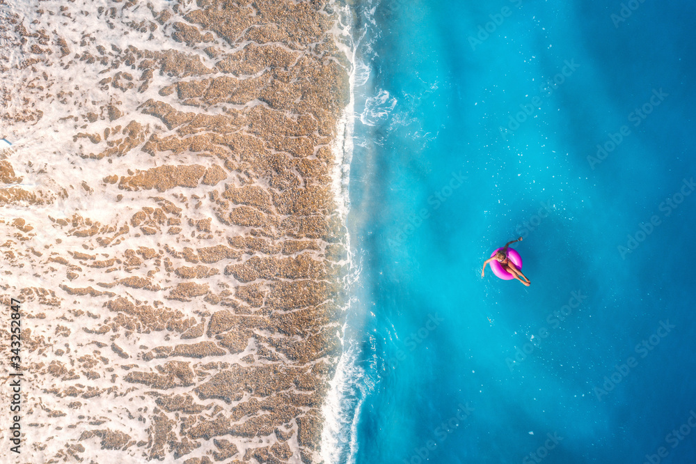 Aerial view of a young woman swimming with pink donut swim ring in clear blue sea with waves at suns