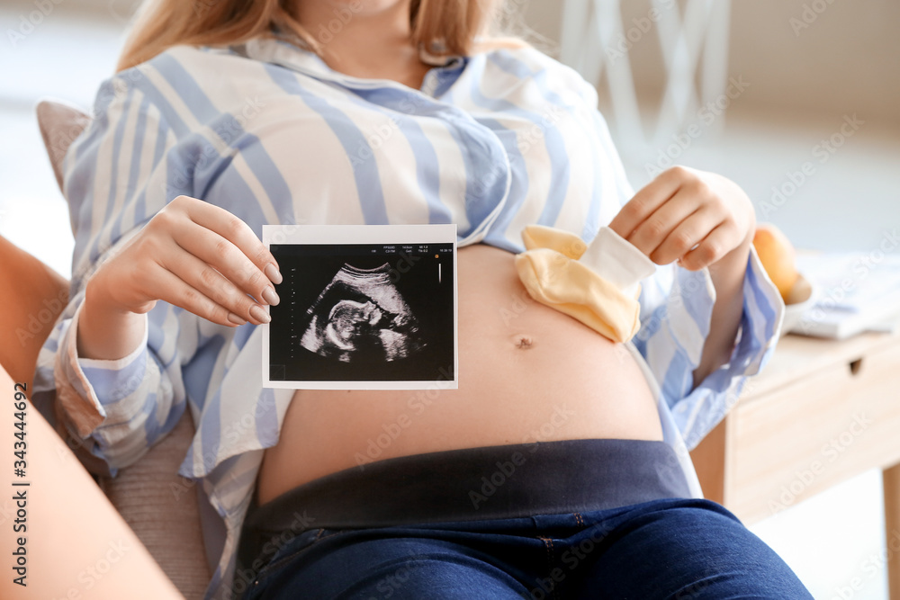 Beautiful young pregnant woman with sonogram image and baby shoes at home