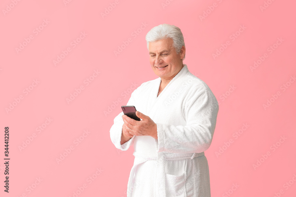 Happy mature man in bathrobe and with mobile phone on color background