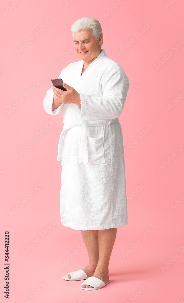 Happy mature man in bathrobe and with mobile phone on color background