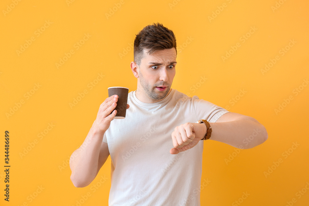 Worried young man with coffee looking at watch against color background