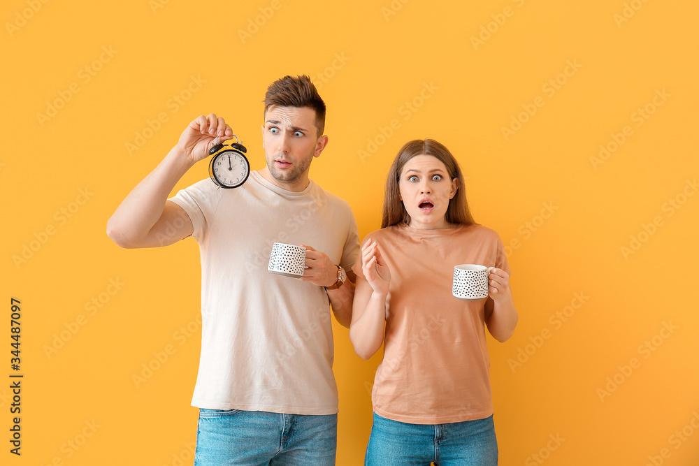 Worried young couple with coffee and alarm clock on color background