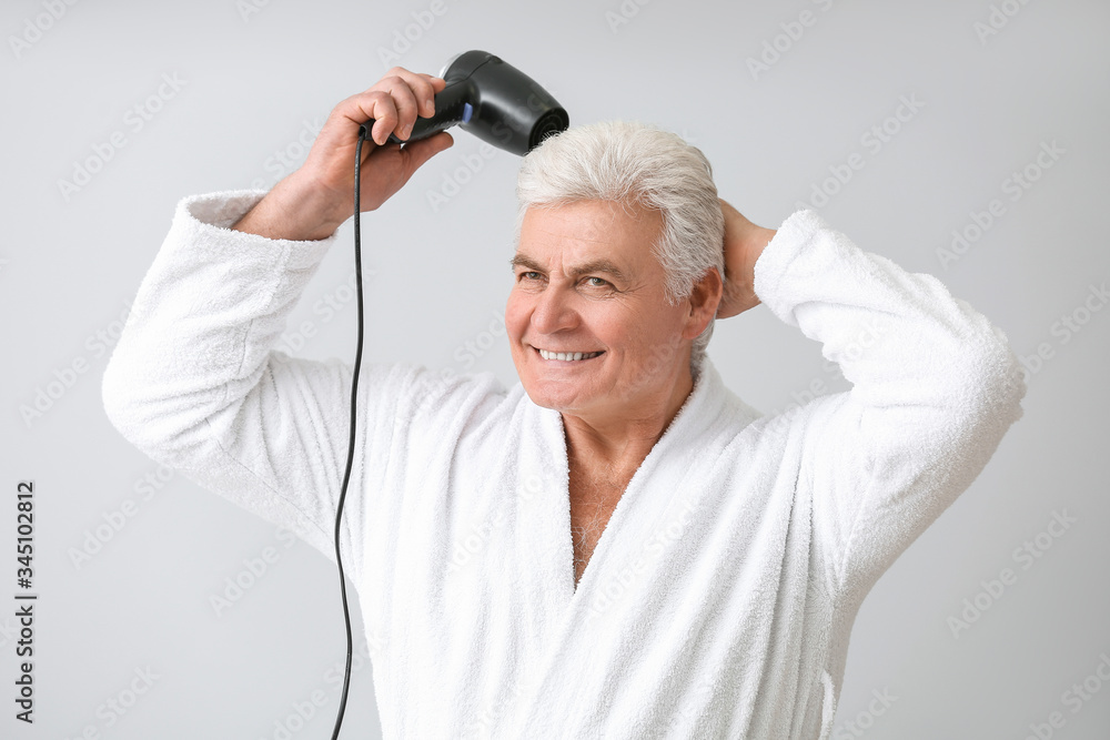 Mature man in bathrobe and with blow dryer on grey background